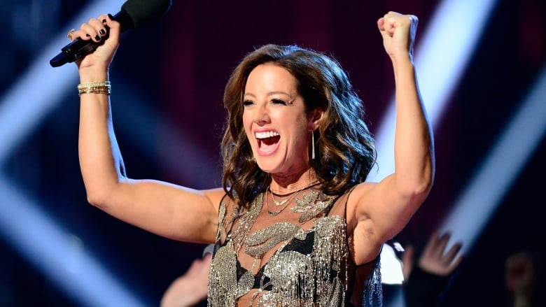 Sarah McLachlan, a white woman with shoulder-length brown hair, raises her arms in celebration while holding a mic.