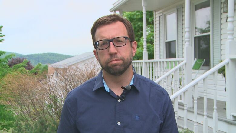 Dark-haired man with beard and glasses standing in front of a white house.