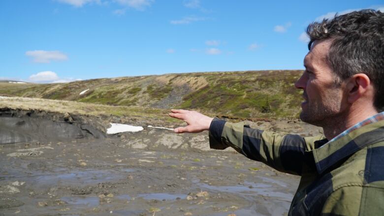 A man in a green coat on the right side of the photo points his right hand out toward a grassy and muddy environment in the background.