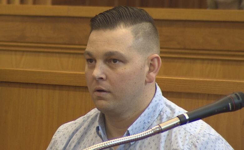 A man is wearing a dress shirt and is sitting in a wooden prisoner's box. His hair is gelled back on top, with the sides closely cropped.