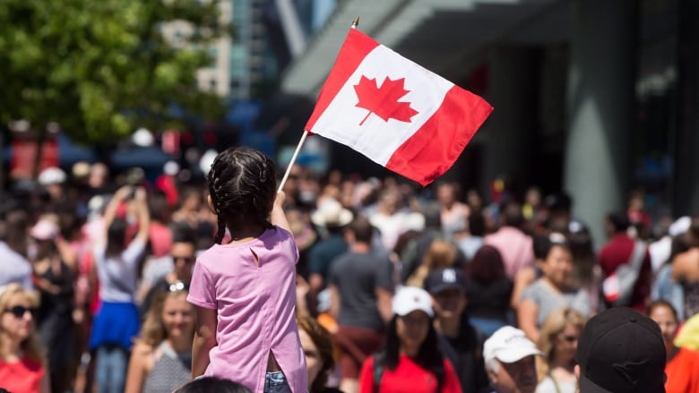 A young girl waves a Canadian flag while being carried on shoulders through a crowd of people.