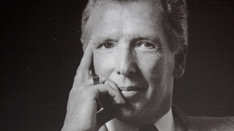 A black and white photo of a man in a suit and tie sitting looking at the camera with his fingers cradling his temple and chin.