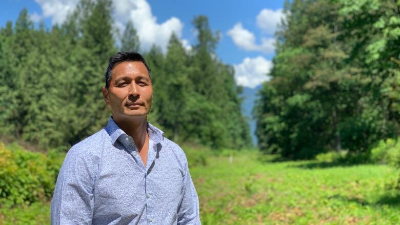 Man stands in a collared shirt in front of a lush green backdrop and blue skies.