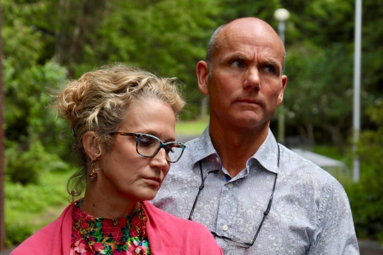 Two parents look sadly at the ground with foliage behind them.