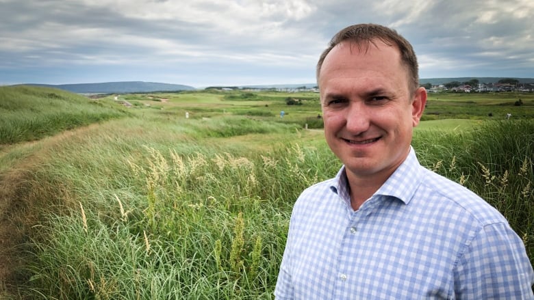 A mman stands in the foreground with dune grasses and a golf course in the background.