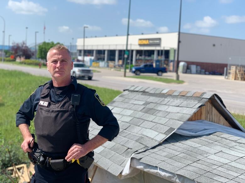 A uniformed police officer stands in front of a wooden shack.