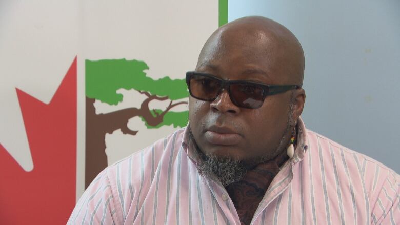 A man wearing sunglasses sits in an office with a logo of a tree on the wall behind him.