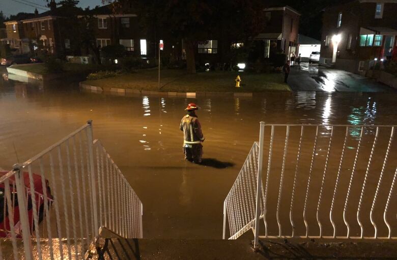 At night, a firefighter stands in the middle of a residential city street, up to his knees in floodwater.