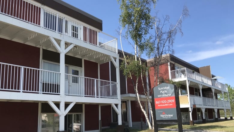 The Sunrdige Place apartment building in summer, with red paint and white balconies and a sign in front.