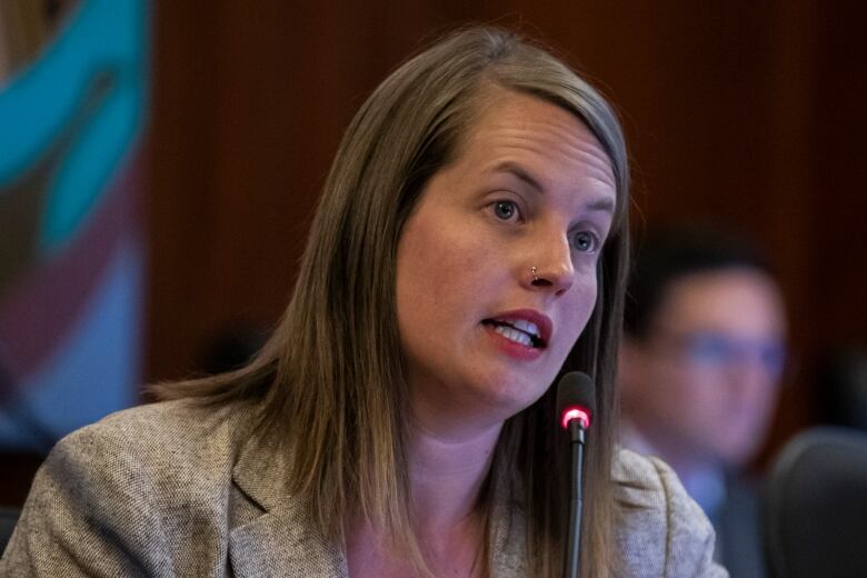 A brown-haired woman with a nose piercing speaks into a mic at council chambers.