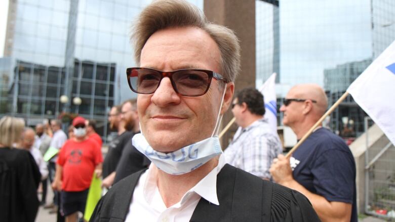 A man with sunglasses and a mask on his chin stands in a crowd. 