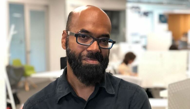 A man with glasses and a beard in a black button up shirt, with collar undone, looks towards the camera in an office space.