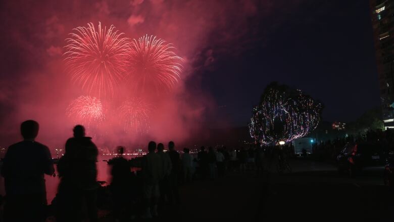 A fireworks display in Vancouver in July 2019. Red-coloured fireworks light up the night sky over the ocean as people in shadow in the foreground look on.