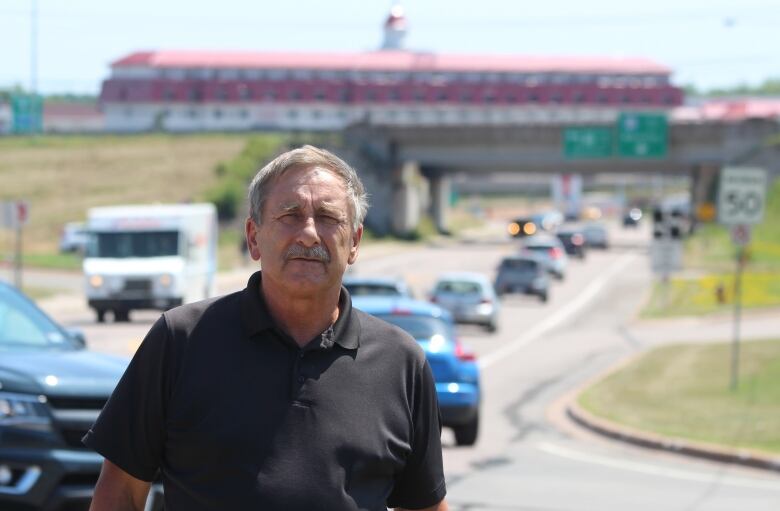 A man with short grey hair standing in front of a busy roadway with a highway overpass and a red-roofed building in the background.