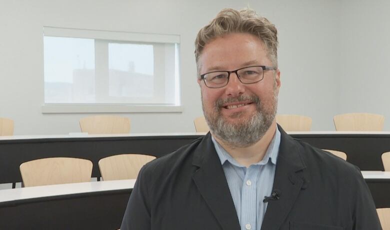 Smiling man with beard and glasses standing in a classroom.