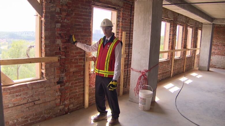 A man wearing a safety vest and hard hat touches a brick building.