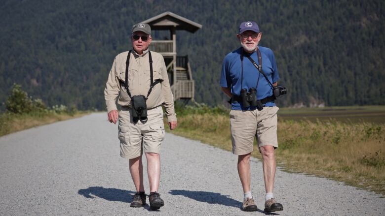 Two men in brown short  and baseball caps with binoculars slung around their necks walk on a park path.