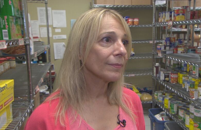 A middle-aged blonde woman is shown in a pink shirt from the shoulders up. She is standing by stocked food shelves.