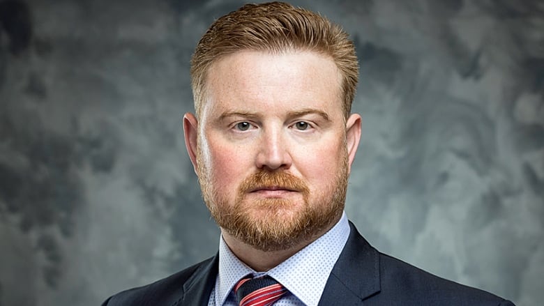 A man with a beard wearing a suit and tie faces the camera.