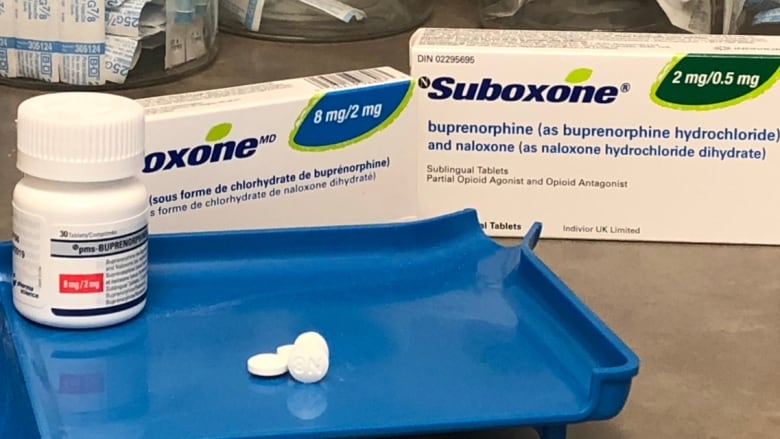 White Suboxone pills are displayed on a blue tray with a bottle and small boxes of the drug in the background.