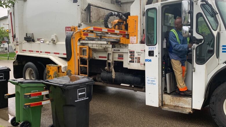 A man dressed in blue stands at the door of a waste collection truck. Waste bins sit along the curb. 