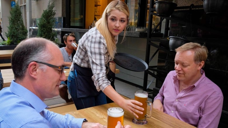 A server places a pint of beer on a table in front of a man. Another man sits across the table holding a pint of beer.