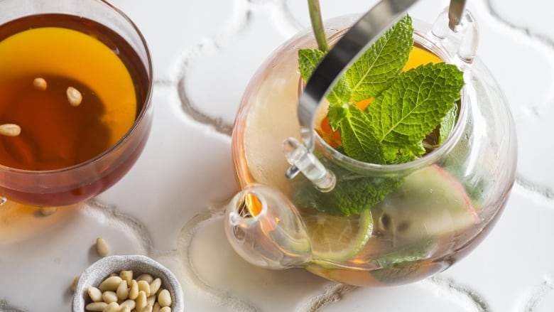 Overhead shot of a glass teapot of mint tea on a white tiled countertop. A cup of tea sits next to it. 