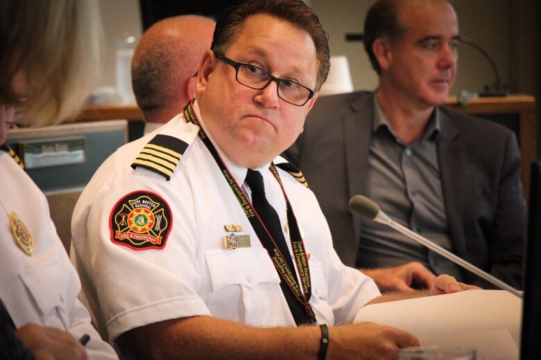 A man wears glasses and a white uniform shirt with a fire department crest on one shoulder.