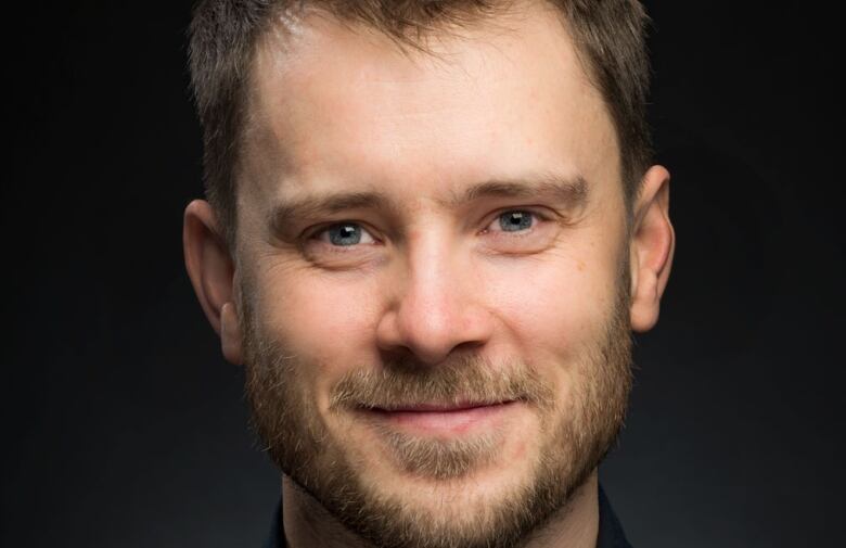 A white man with a beard against a dark background. 