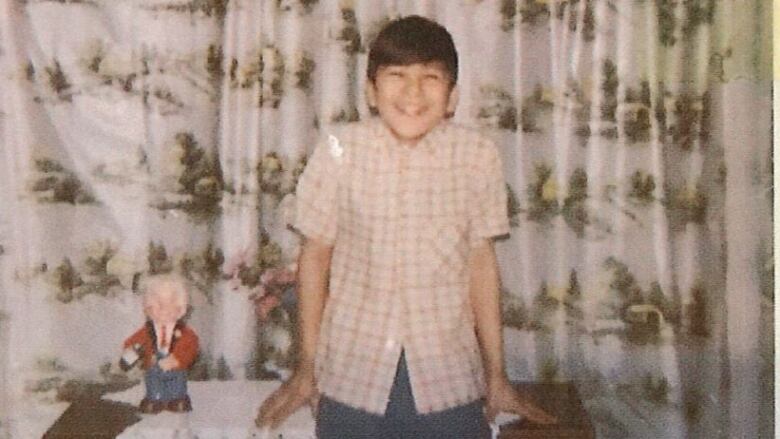 A 1960s photo of a teen boy standing in front of furniture and a curtain.