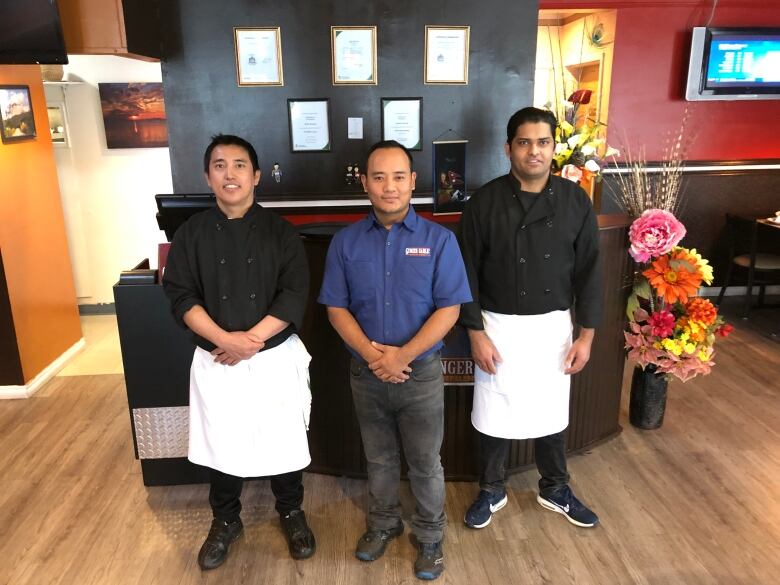 Three men pose in a restaurant. Two are wearing chef's jackets.