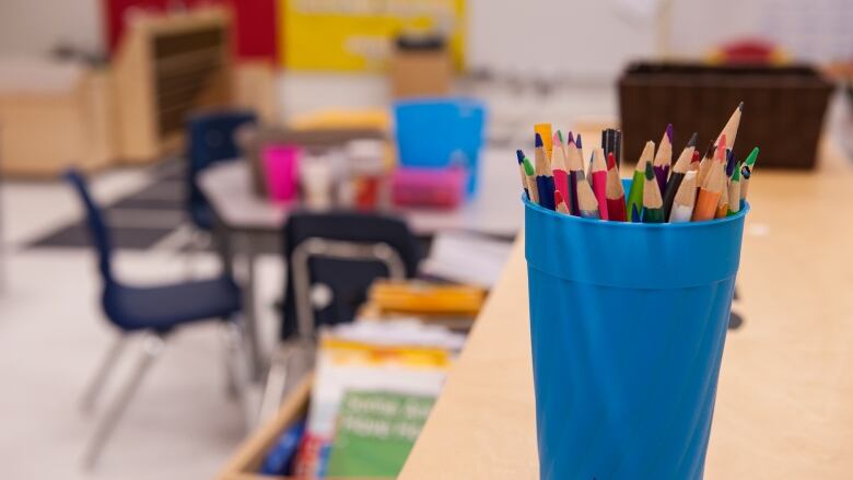 Coloured-pencils are in a bright blue cup. It sits on a table in a classroom. 