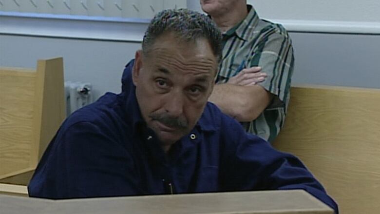 A person with short grey hair and a grey moustache sits in a courtroom wearing a dark blue long sleeve shirt.