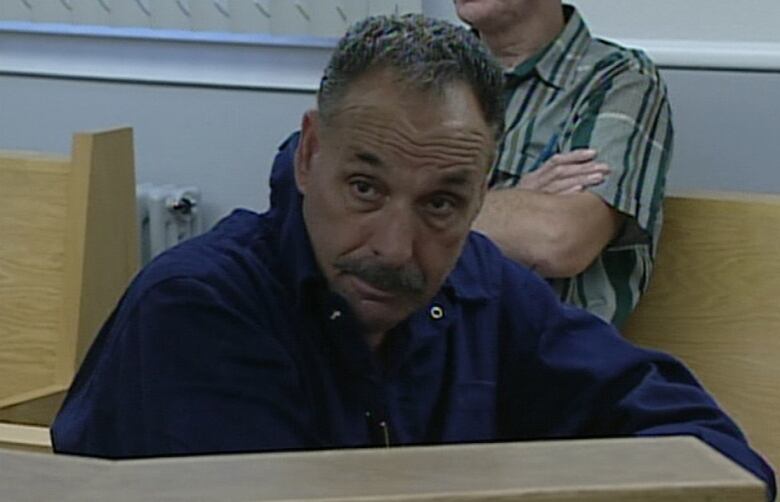 A person with short grey hair and a grey moustache sits in a courtroom wearing a dark blue long sleeve shirt.