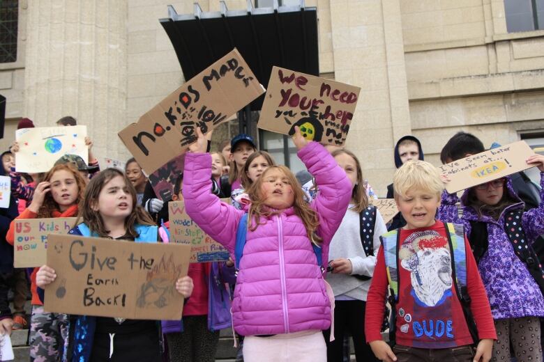 Elementary-aged students in bright clothing stand at  the Manitoba Legislature holding signs with messages on climate change.