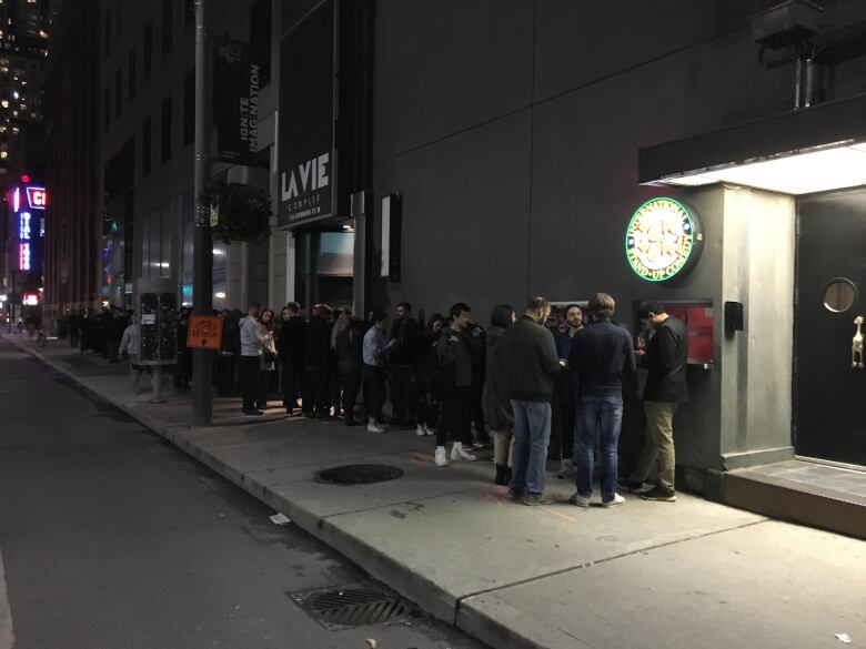 A crowd lines up outside a club at night on a city street. The lineup stretches one city block.