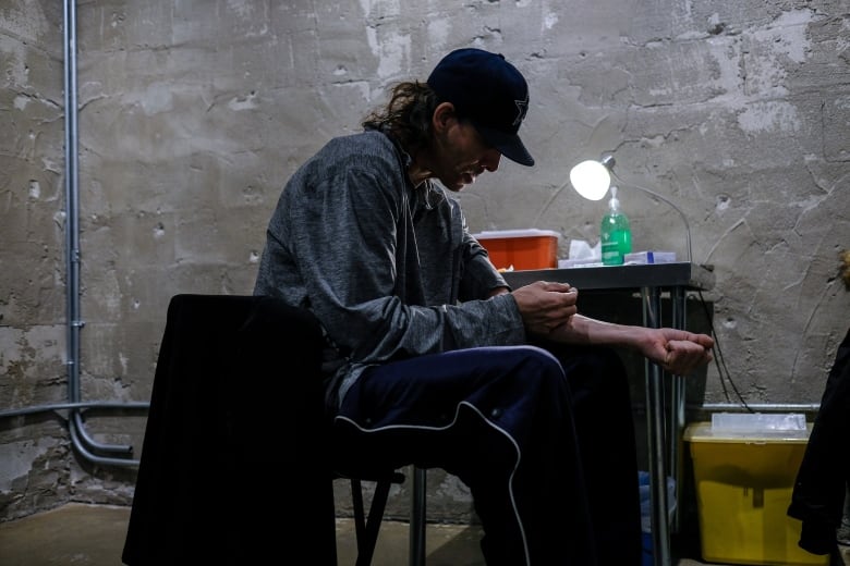 A drug user wearing a hat sits in a chair near a table with a lamp and injects a needle into their arm.