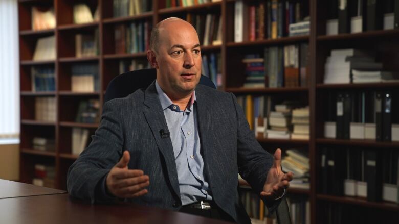 A portrait of a man in a dark blue suit jacket behind a bookcase. 