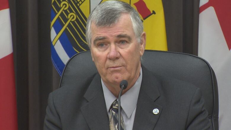 A man in a suit sits in front of a microphone with flags behind him.