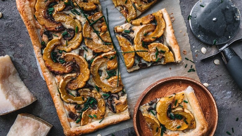 A rectangular tart with slices of delicata squash on it sitting on a dark grey surface. A slice of the tart is sitting on a small wooden plate. 