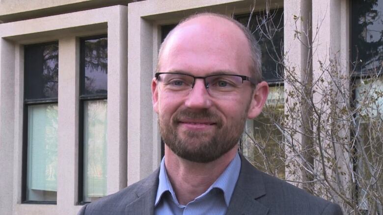  A man wearing glasses stands in front of a grey building.