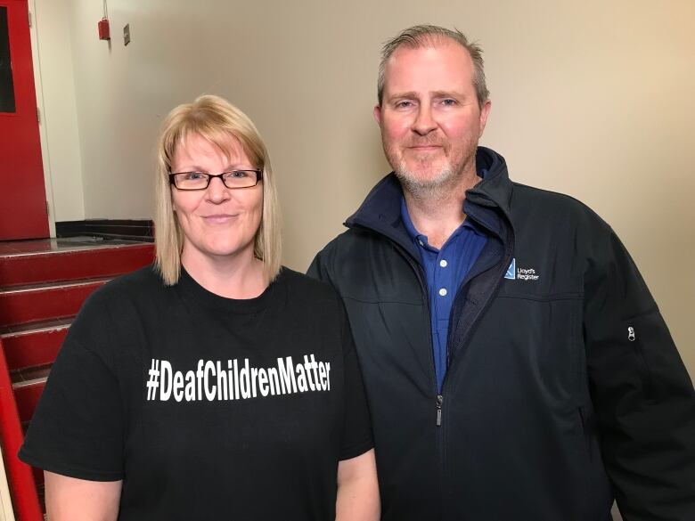 A blonde woman standing next to a grey-haired man. She's wearing a black T-shirt that says Deaf Children Matter. He's wearing a navy blue windbreaker over a royal blue buttom-up shirt.