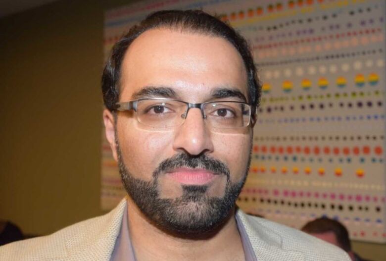 A man with brown hair and a beard, and wearing glasses, stands near a colourful wall hanging.