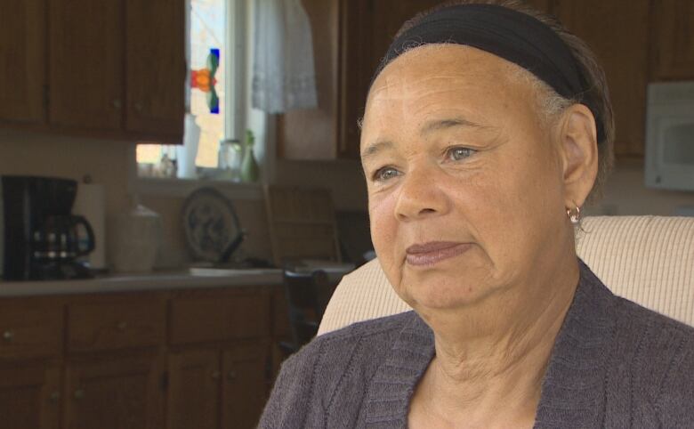 A Black woman with a black headband sits in a chair with a kitchen behind her