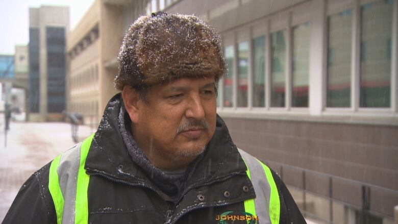 A man wearing a fur hat with snow on it and a jacket with yellow reflective stripes stands on a sidewalk, looking to the right of the camera.