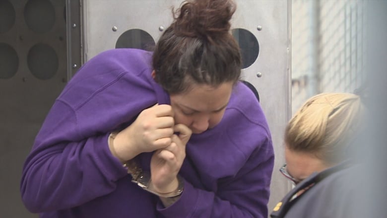 A white woman has her dark hair in a bun. She is wearing a purple sweater and her hands are in handcuffs. She is trying to use her sweater to block her face