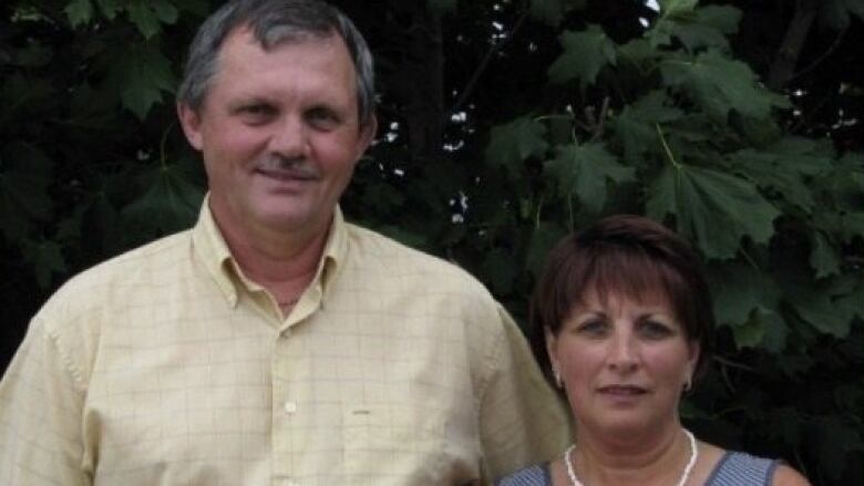 A tall grey-haired man in a yellow shirt smiles. He is standing next to a much-shorter woman with cropped dark hair. 