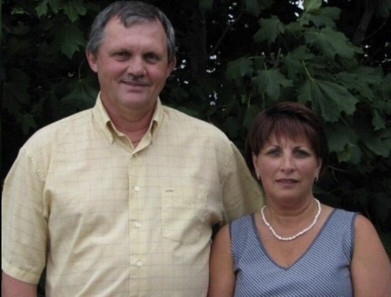 A tall grey-haired man in a yellow shirt smiles. He is standing next to a much-shorter woman with cropped dark hair. 