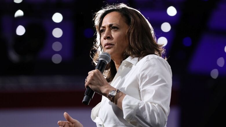 A woman in a white suit speaks into a microphone.