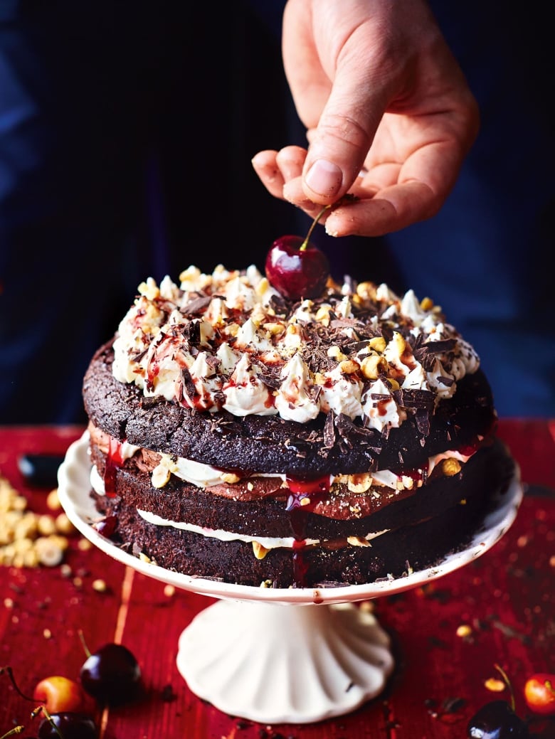 3-layer black forest cake topped with Chantilly cream, cherry pie filling, chocolate and cherries, sitting on a white cake pedestal. 